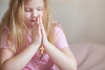 A blonde girl child prays at home in the morning with her hands folded with a cross in front of her.  Religion, Christianity, faith in God concept
