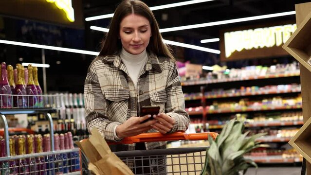 Pretty young woman uses smartphone while walking with a shopping cart in a supermarket