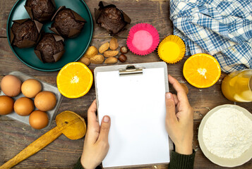 glass bottle of fresh orange juice with fresh fruits and almond, hazelnuts and homemade chocolate muffins and flour, hands woman holding a empty clipboard for your recipe on wooden table