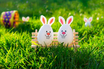 Creative Easter composition of Funny Easter eggs with bunny ears sitting on bench on green grass background with fallen basket. Sunny festive mood. Fairy tale Easter hunt. Selective focus. Copy space