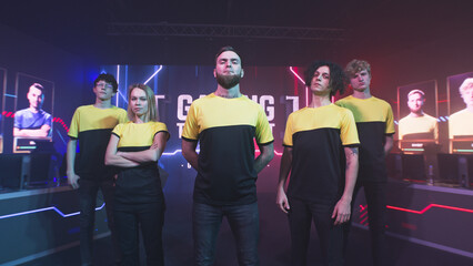 Assured team of teammates esportsman gamers in yellow uniform standing near bearded team leader and looking at camera during professional gaming competition