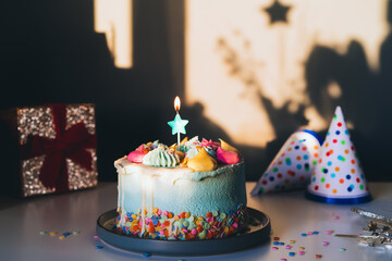 Colorful birthday cake with sprinkles and star shaped candle, festive caps and gift box on a dark wall background with shadows. Selective focus, copy space.