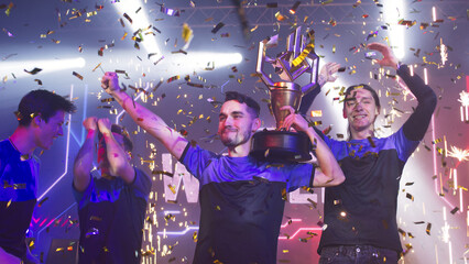 Esport gamers team with golden trophy celebrating victory amidst confetti and pyrotechnics after professional esports competition