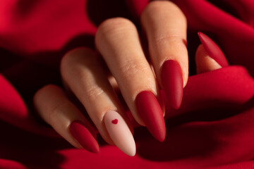 Matte red nails with small red heart on beige colour nail on the red fabric background. Saint Valentine's nail design. 