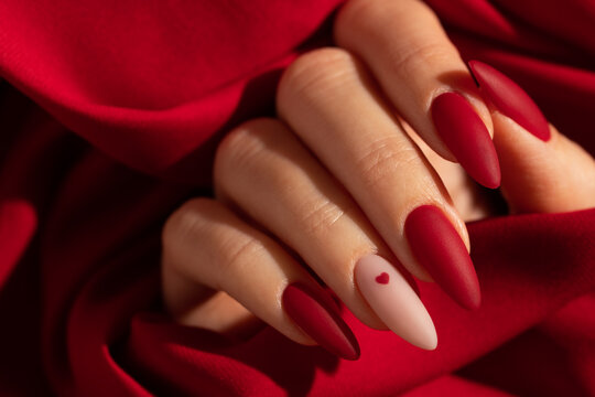 Matte red nails with small red heart on beige colour nail on the red fabric background. Saint Valentine's nail design. 