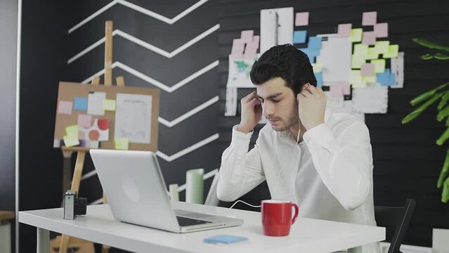 A young man of Caucasian appearance is sitting at a computer and working remotely. The young man puts on headphones and communicates via video link