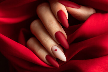 Matte red nails with small red heart on beige colour nail on the red fabric background. Saint Valentine's nail design.  - obrazy, fototapety, plakaty
