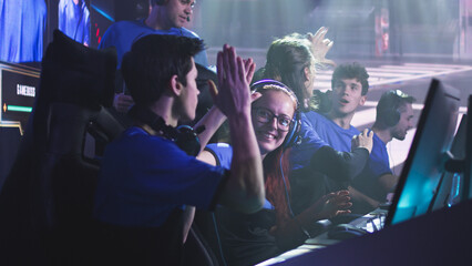 Cheerful young woman gamer esportsman in glasses and headphones smiling and giving high five to male teammates while celebrating victory in video game match during professional tournament - obrazy, fototapety, plakaty