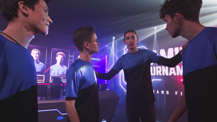 Esportsman gamers teammates in blue uniform stacking hands together then taking seat at desk with computers at start of gaming tournament