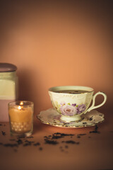 a white and pink cup with flowers with black tea and a ceramic tea jar and scattered tea and a burning pink candle in a glass with pearls on a pink background