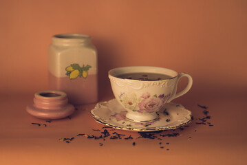 Obraz na płótnie Canvas a white and pink cup with flowers with black tea and a ceramic tea jar and scattered tea on a pink background 