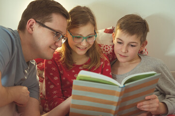 Young father reading book with his children in bed