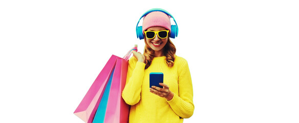 Colorful portrait of stylish smiling young woman listening to music in headphones with shopping bags posing wearing a yellow knitted sweater, pink hat isolated on white background