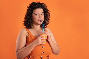 Woman drinking fresh beverage, curly hair, wearing an orange outfit, holding an orange plastic cup with a blue plastic straw, looking at the camera, by an orange background as a negative space.