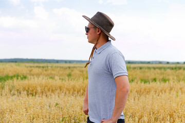 Naklejka na ściany i meble Crop and harvest. Portrait of farmer standing in gold wheat field with blue sky in background. Young man wearing sunglasses and cowboy hat in a field examining wheat crop. Oats analysis