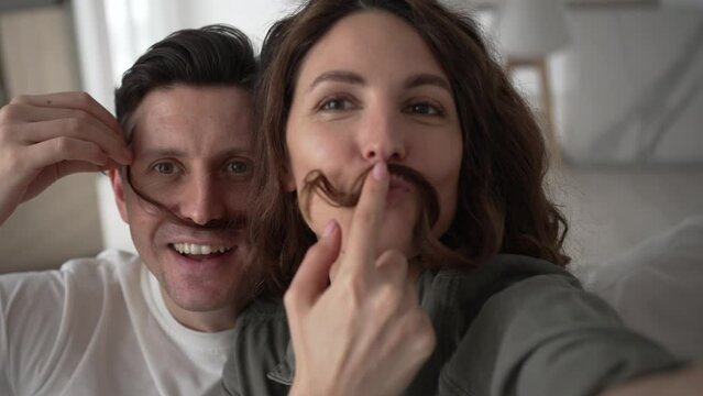 Happy newlyweds play and pose making mustaches with curly brunette hair and kiss smiling broadly for selfies via smartphone closeup