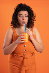 Woman drinking fresh beverage, curly hair, wearing an orange outfit, holding an orange plastic cup with a blue plastic straw, looking down at the cup, by an orange background as a negative space.