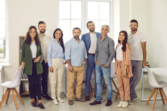 Group Portrait Of Happy Confident Business Professionals And Teammates At Work. Team Of Young And Mature People In Smart Casual Clothes All Together Standing In Office, Looking At Camera And Smiling