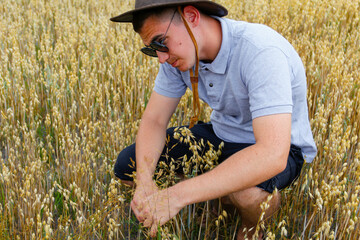 Crop grain. Portrait of farmer seating in gold wheat field with blue sky in background. Young man wearing sunglasses and cowboy hat in field examining wheat crop. Oats grain industry. Closeup