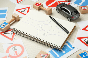 Many different road signs, notebook with sketch of roundabout and toy car on white wooden background. Driving school
