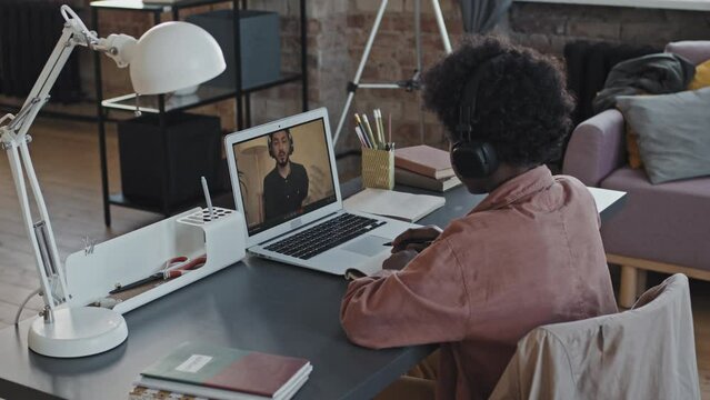 Over-shoulder of elementary-aged Black boy wearing over-ear headphones, sitting at desk at home in afternoon, having online lesson with Asian male teacher on portable computer