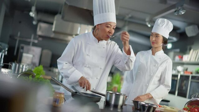 Restaurant Kitchen: Portrait of Asian and Black Female Chefs Preparing Dish, Tasting Food, Doing High-Five in Successful Celebration. Two Professionals Cooking Delicious, Authentic Food, Healthy Meals
