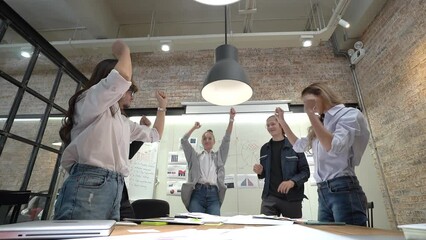 Low-angle view of a young successful business team, Caucasian and Asian, men and women standing around table with many paper sheets, jumping and dancing happily after completion of a project in office