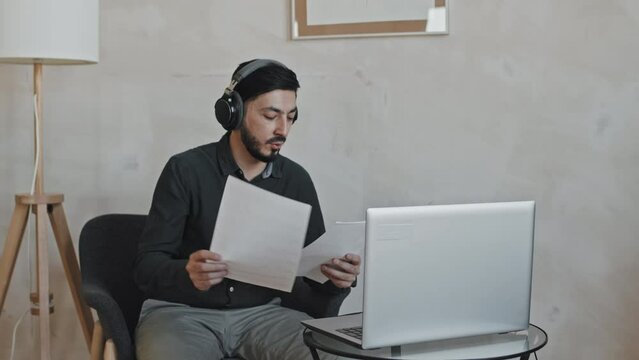 Medium long of Asian teacher wearing beard and over-ear headphones sitting in armchair at home in afternoon, teaching online lesson using portable computer