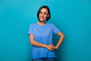 Close up portrait of happy beautiful young brunette woman with stylish hairstyle in blue t-shirt while she is posing and isolated on blue background in studio