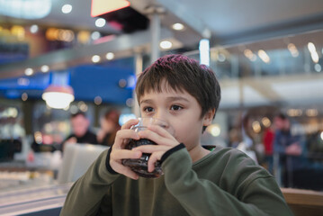 Portrait of  Young kid sitting on table drinking cold drink in restaurant, A boy drinking soda or soft drink from glass, Child boy waiting for food in cafe