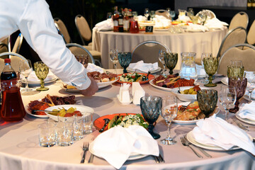 Table is set with variety of gourmet dishes and appetizers for banquet. Waiter's hand putting plate on table. Selective focus.