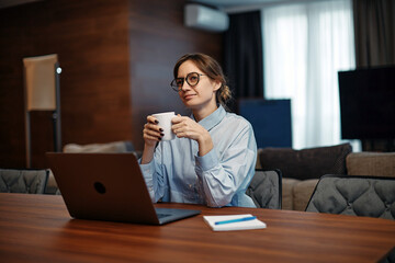Smiling woman employee sit at desk with laptop at home office hold coffee tea cup enjoy morning, dreaming. Distant job