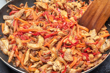 A wooden spatula mixes pork meat in a pan with peppers, carrots, onions.