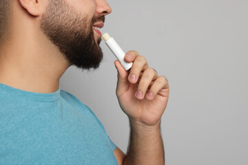 Young man applying lip balm on grey background, closeup
