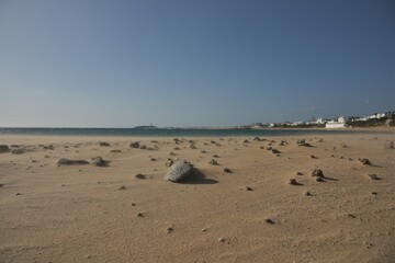 Miniature sandstorm creates a windswept strewn stone and pebble moonscape at Playa del Pirata yellow sand beach with Canos de Meca township and Cabo de Trafalgar lighthouse in the background