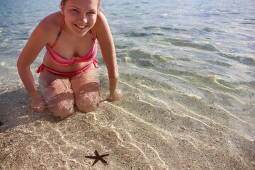Happy face of pretty sunburn blonde teen girl which finding red starfish on the shallow beach of tropical island. Year round traveling and beach family vacations.