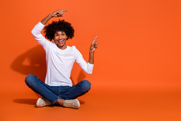 Photo of attractive excited guy wear white shirt pointing empty space sitting floor isolated orange color background