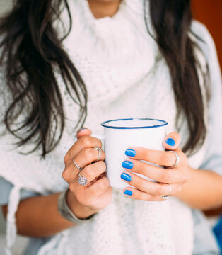 Close view of the hands of a young woman with blue painted nails and finger rings holding a white enameled mug with coffee or tea