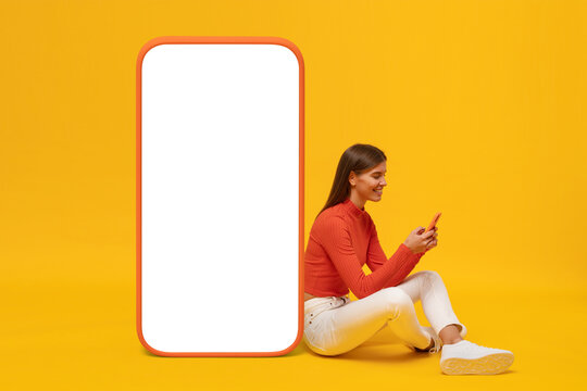 Young woman sitting on floor near huge phone mock up for app