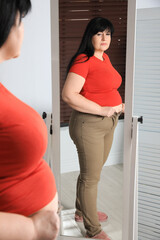 Overweight woman trying to button up tight trousers in front of mirror at home