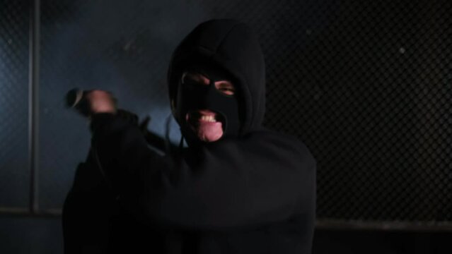 Close up. An aggressive man in a balaclava and hood hits a baseball bat in front of the camera. Assault, beating, fight