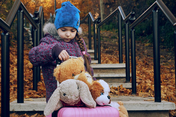 A girl child holds in her hands a pink suitcase full of soft toys. On the steps, a girl tries to...