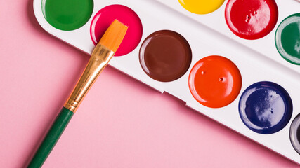 Multicolored watercolor paints with a brush on a pink background. Objects for art