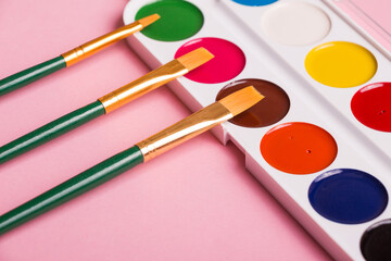 Multicolored watercolor paints with a brush on a pink background. Objects for art
