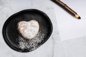 a heart-shaped donut on a plate, sprinkled with sugar, next to a blank sheet with a pencil