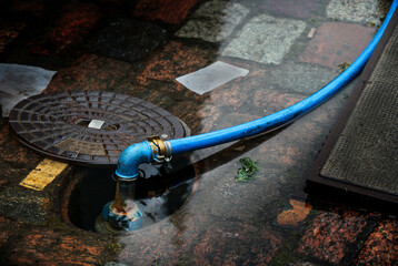 blue water hose connected to a flooded underground hydrant in a cobblestone street, copy space,...