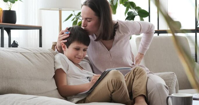 Loving mother kissing son while helping him with homework on sofa at home