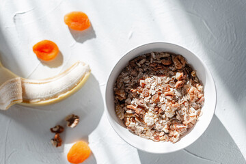 Healthy raw food muesli with oatmeal, dried apricots, walnuts and banana on a white table with early morning sunlight, top view. Vegan breakfast