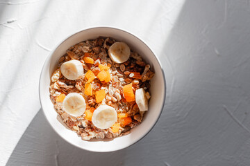 Delicious healthy cheese vegan breakfast, muesli with oatmeal, dried apricots, banana and nuts in a white bowl on a white plate with rays of sun in the early morning