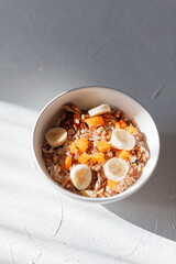 Delicious lactose-free breakfast muesli with oatmeal, dried apricots and banana in a white bowl on a white table at sunrise with shadows.